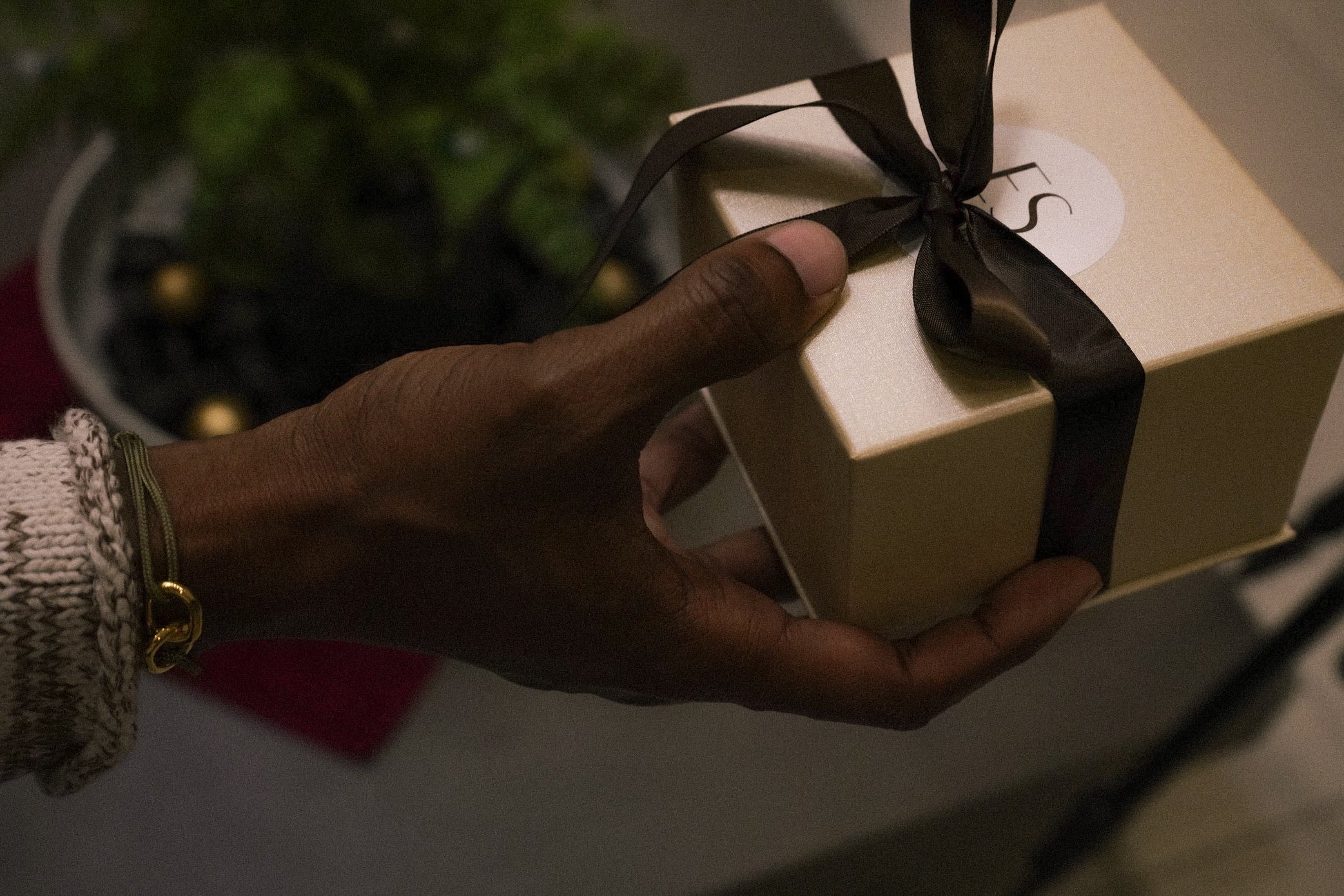 Hand holding a gold gift box with a black ribbon against a blurred indoor background